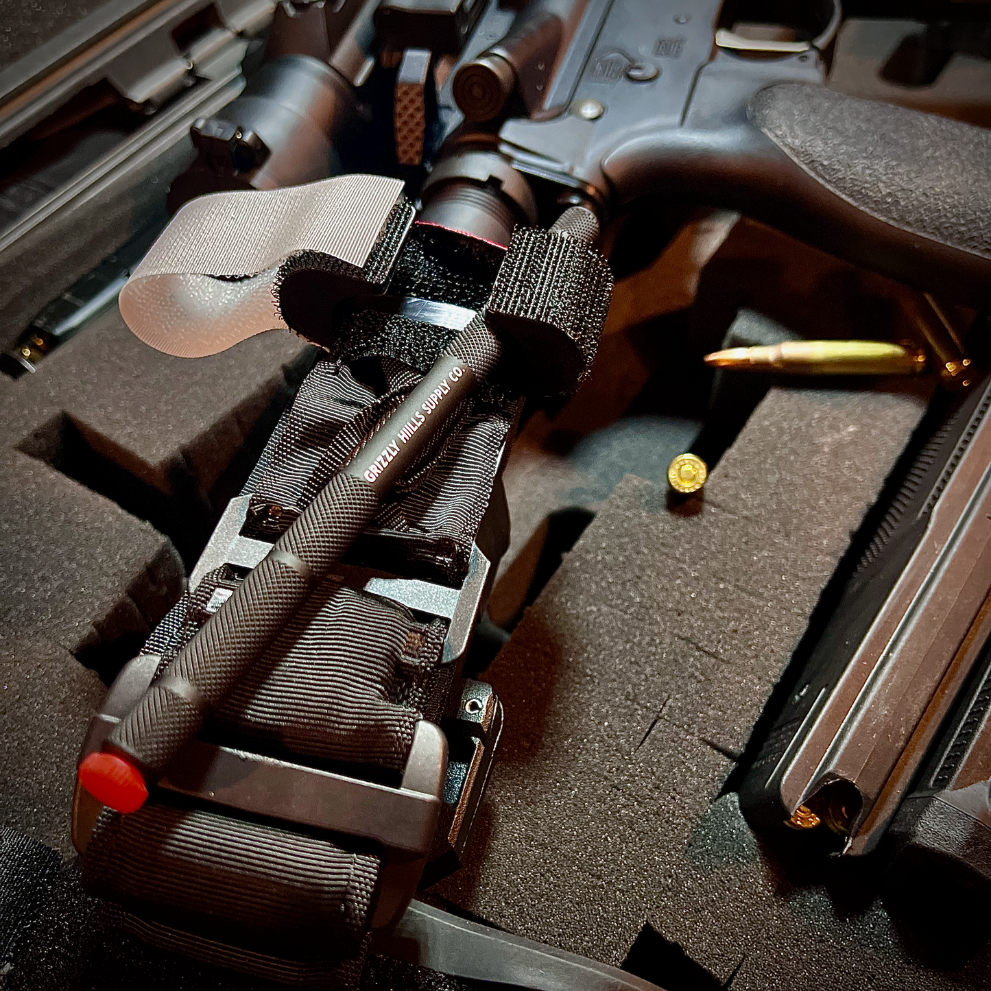 Different shot angle of the tourniquet attached to the stock of an AR-15 rifle with spare bullets and magazines in the background